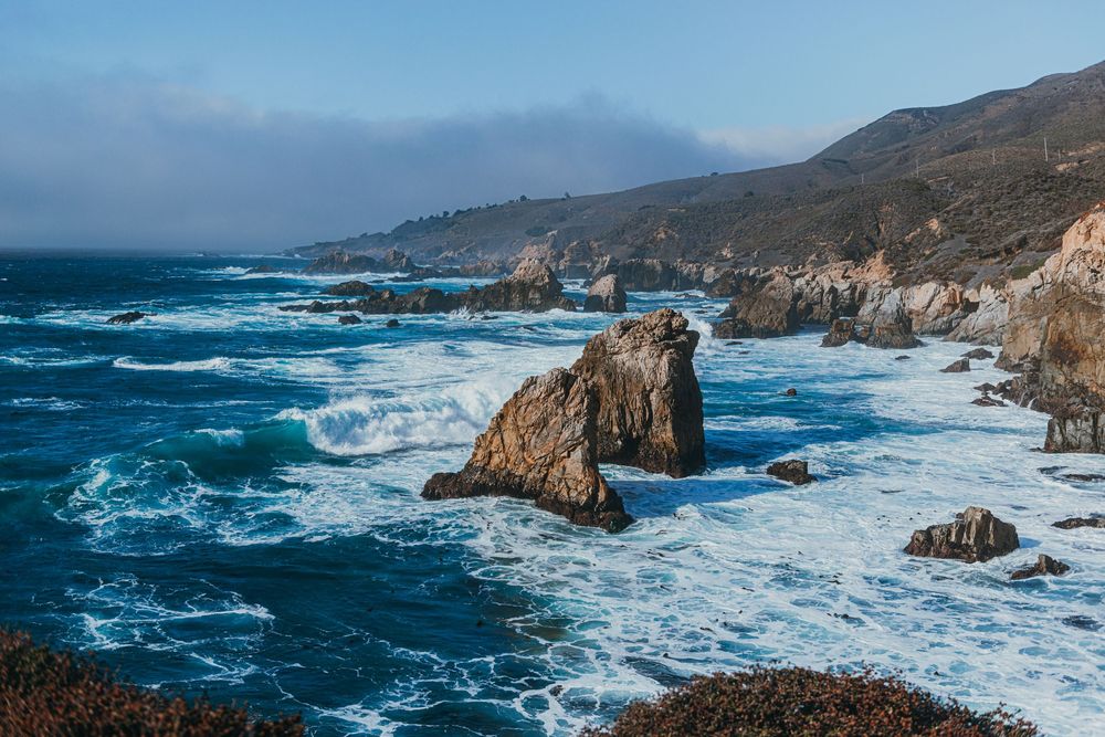 Dramatic panoramic view of rugged Big Sur coastline with crashing waves against rocky cliffs under cloudy skies.