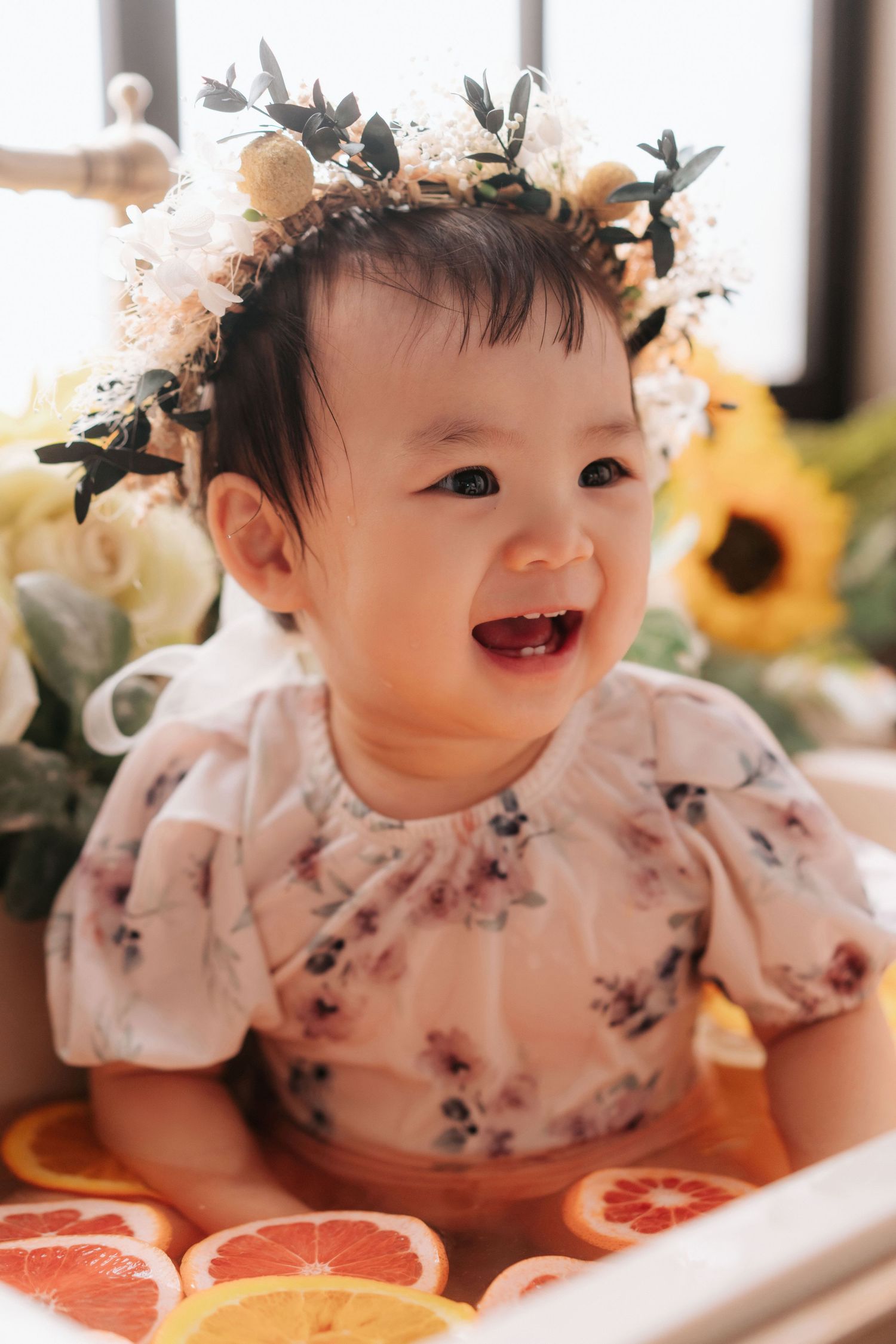 A baby in a floral dress and flower crown smiles while lying on a surface with citrus fruits.