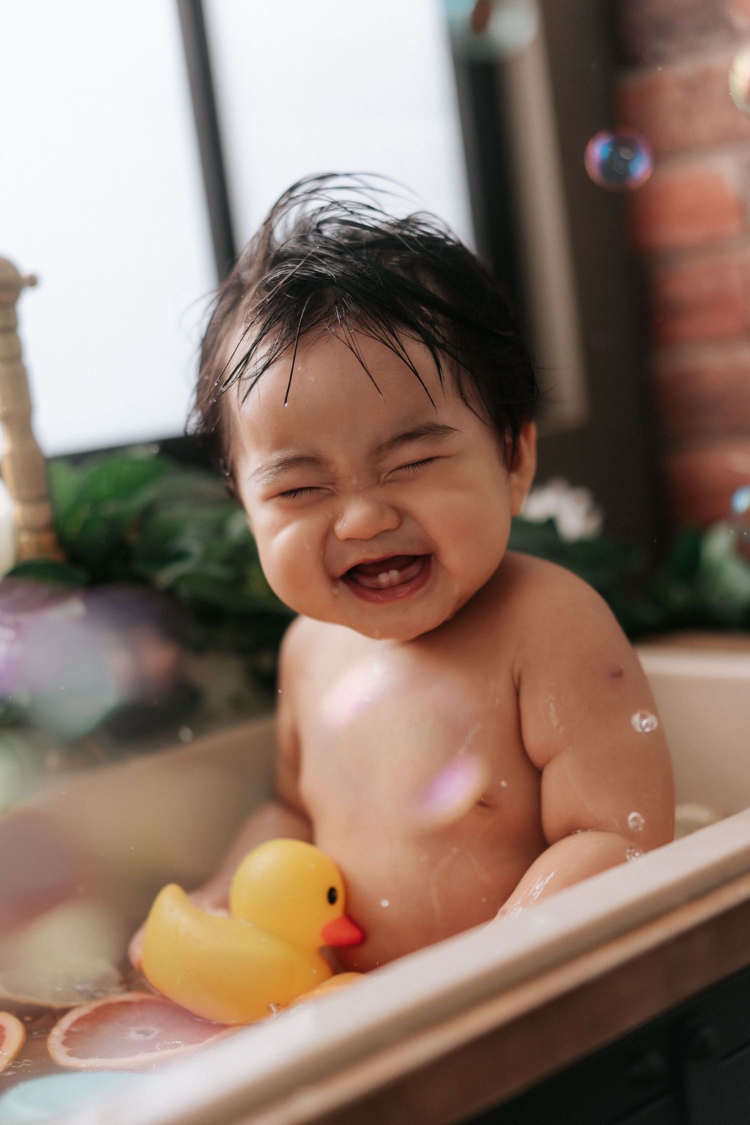 Happy baby laughs during bath time with yellow rubber duck toy in bathtub.
