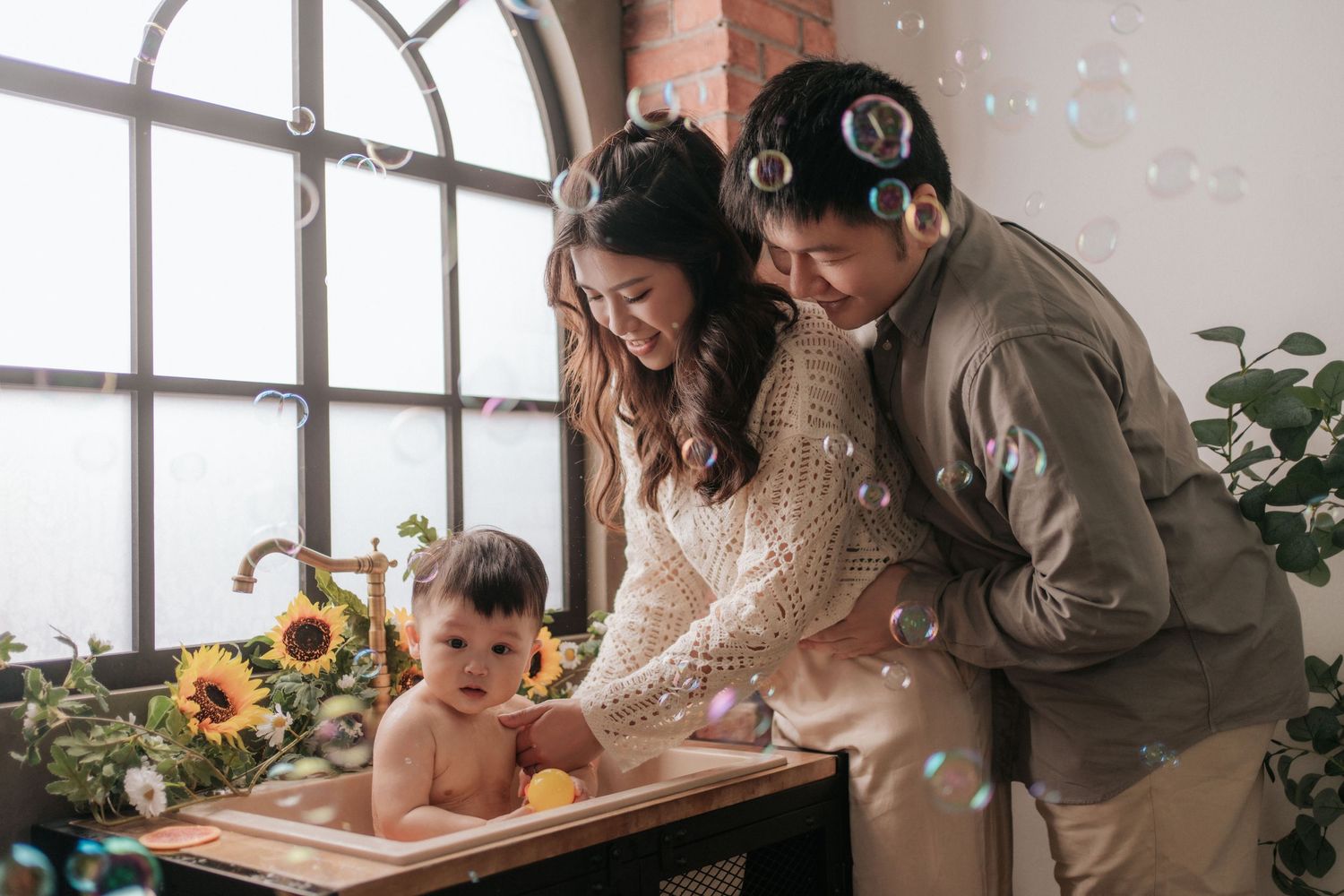 Family members gather around a wooden table in a sunlit room with large windows and plants for a heartfelt moment together.
