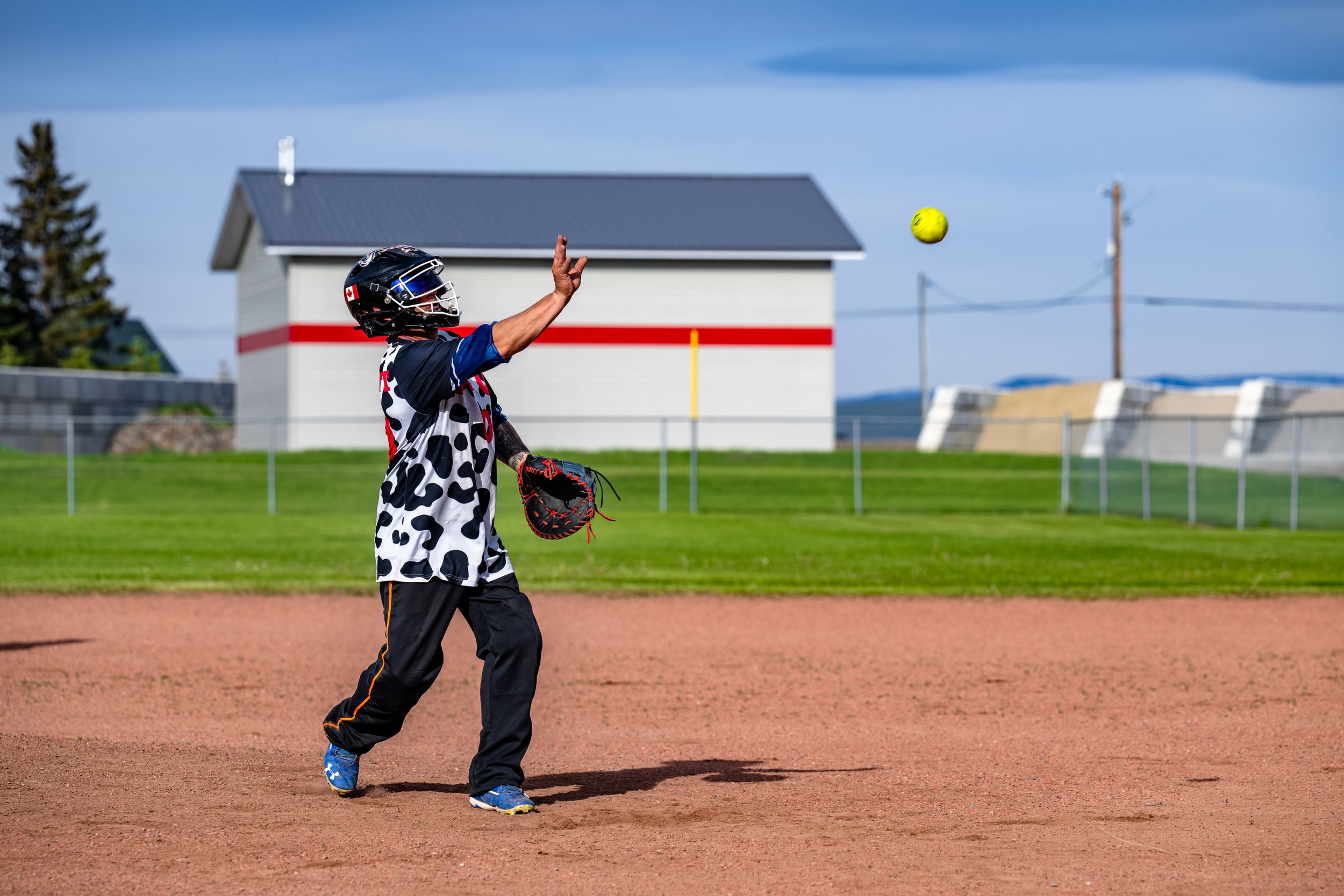 Softball player wearing black and white uniform swings bat at yellow ball on dirt field with white building in background.