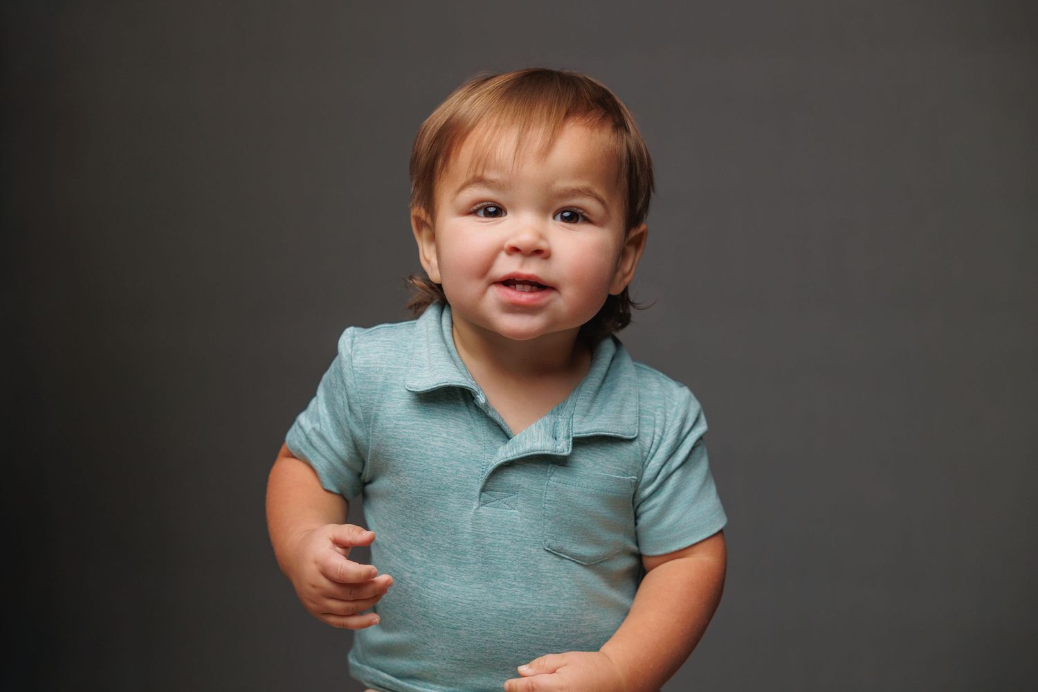 A toddler wearing a teal polo shirt smiles while sitting against a dark background.