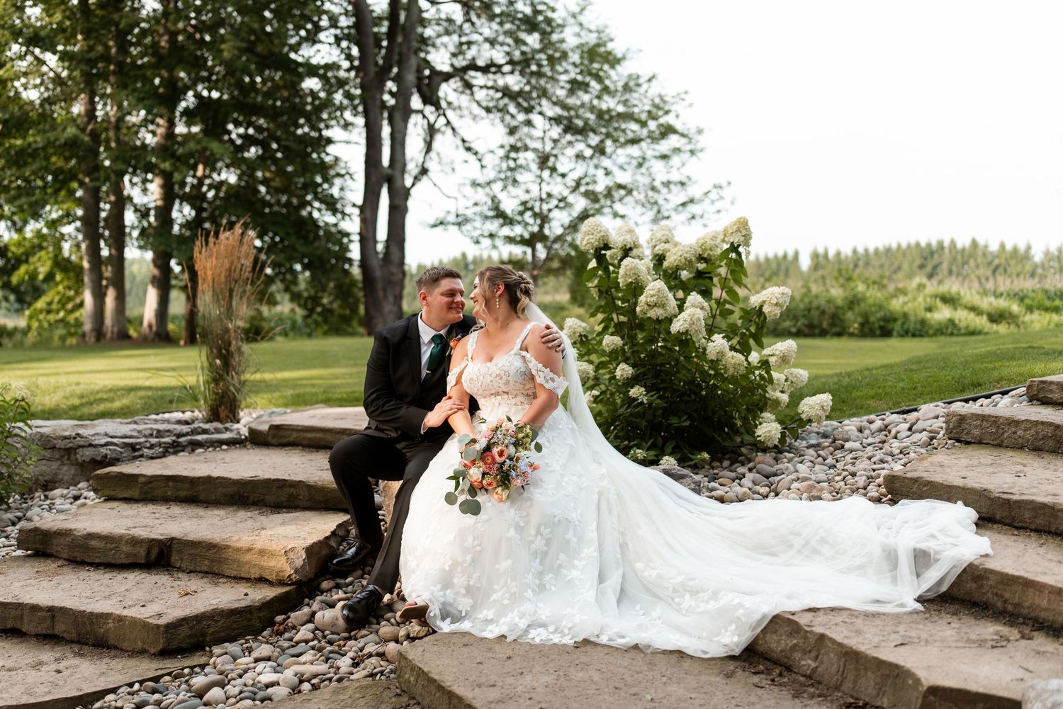 A wedding couple walks together across a scenic park lawn with a flowing white dress train trailing behind them.