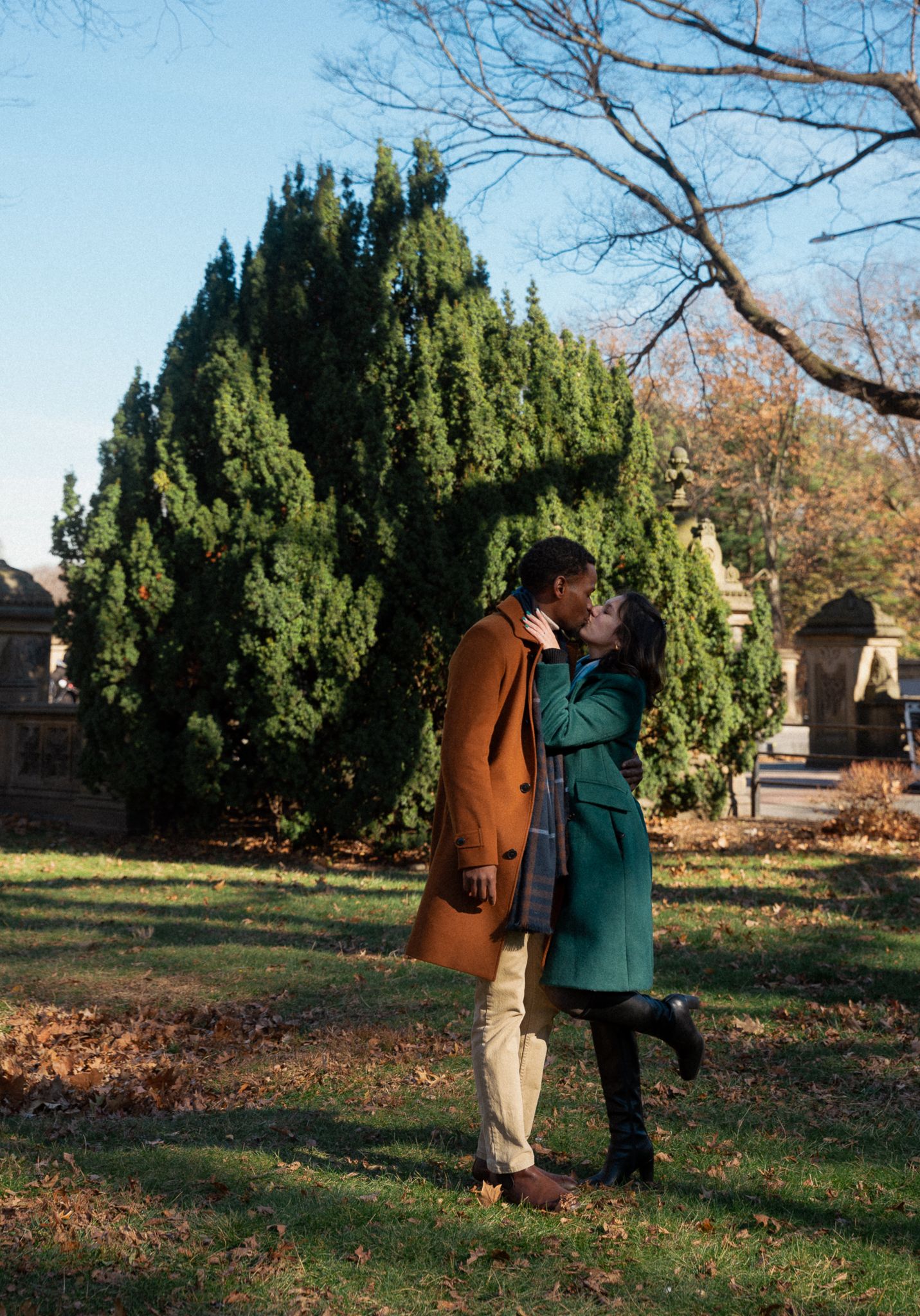 A romantic moment between two people in winter coats near evergreen trees in a park.