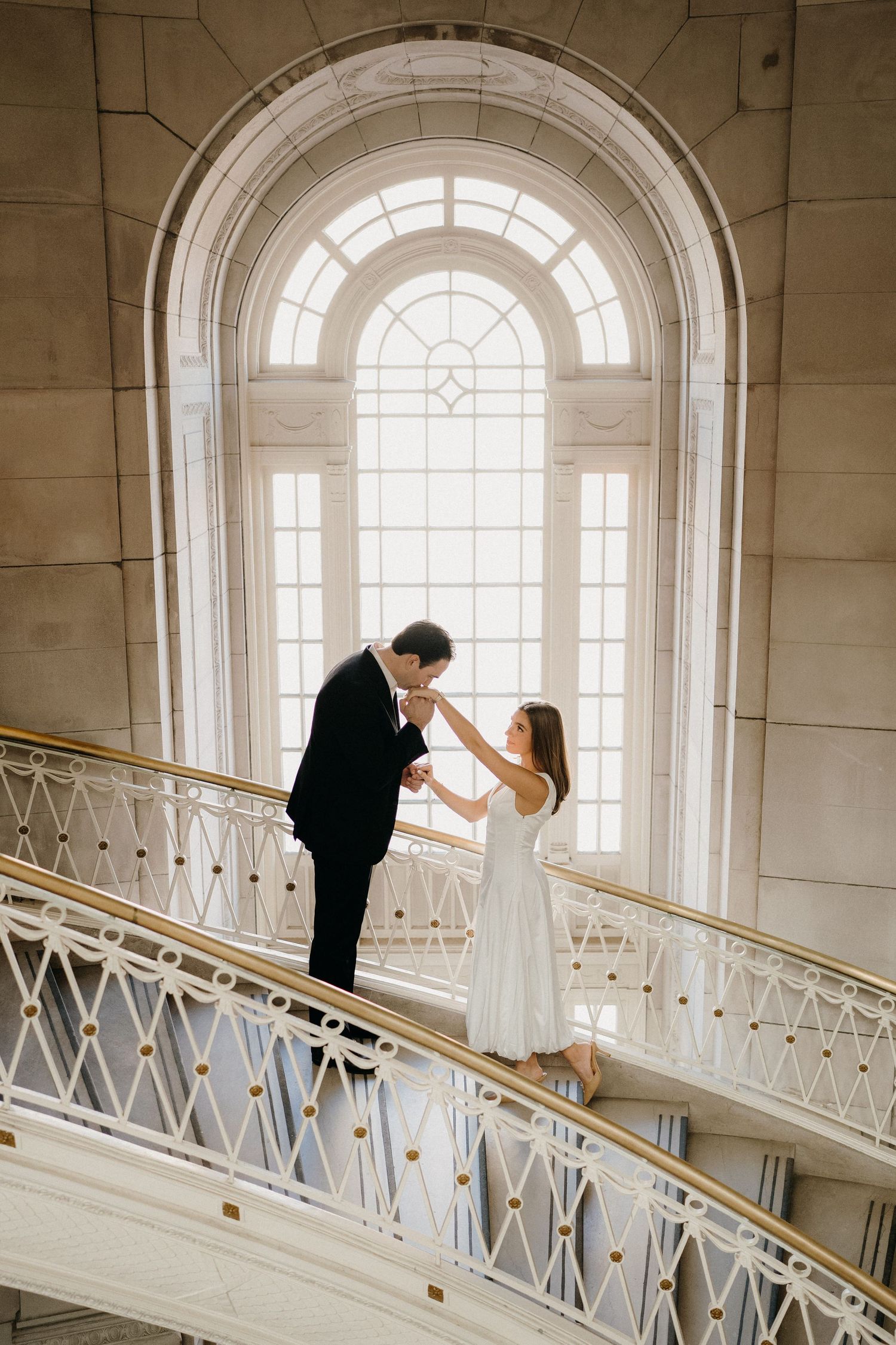 A romantic couple shares an intimate moment on an elegant curved staircase beneath a grand arched window.
