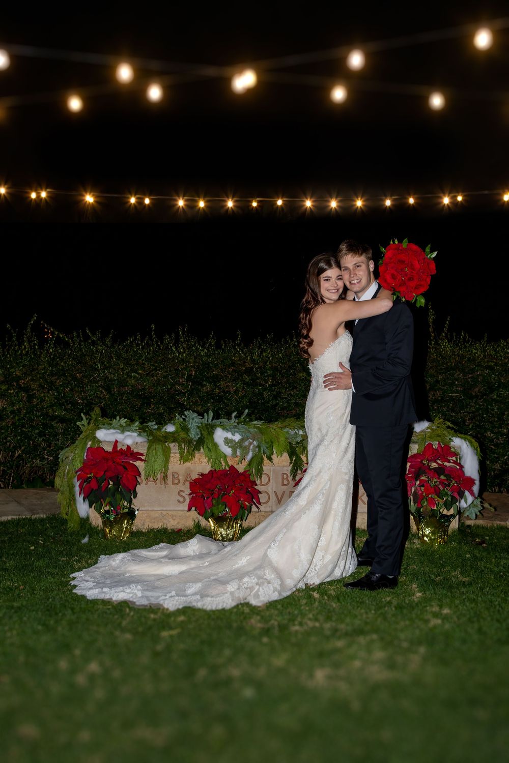 A couple embraces under string lights at night with red poinsettias and holiday decorations on the lawn.
