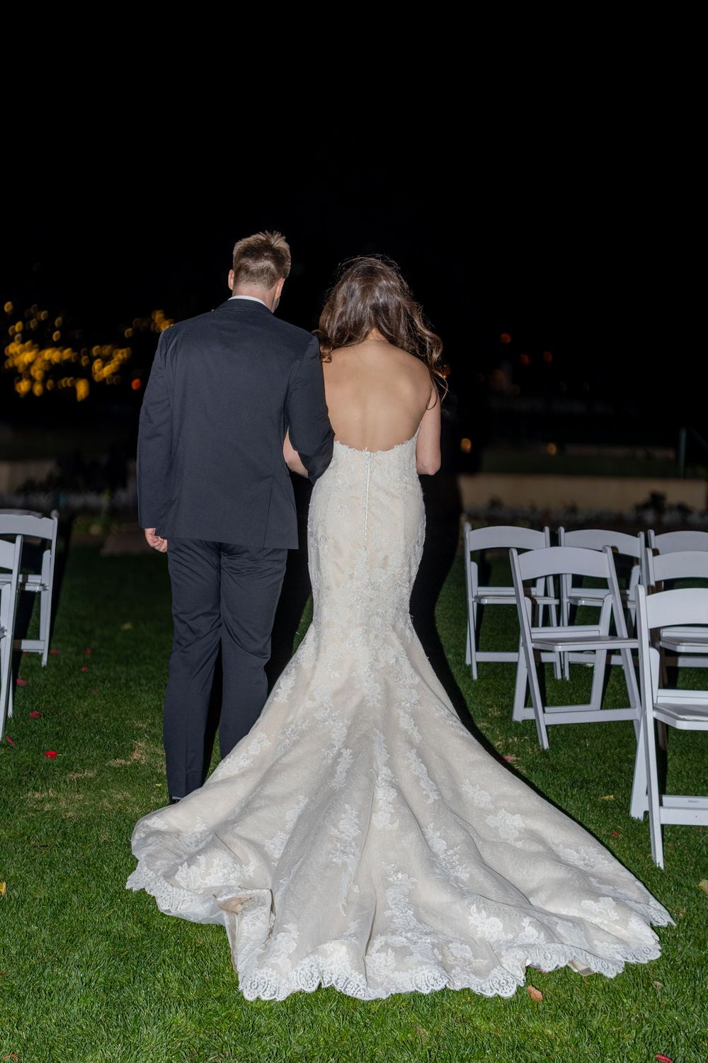 A bride in a white lace wedding dress sits on grass next to rows of white chairs at an outdoor evening ceremony.