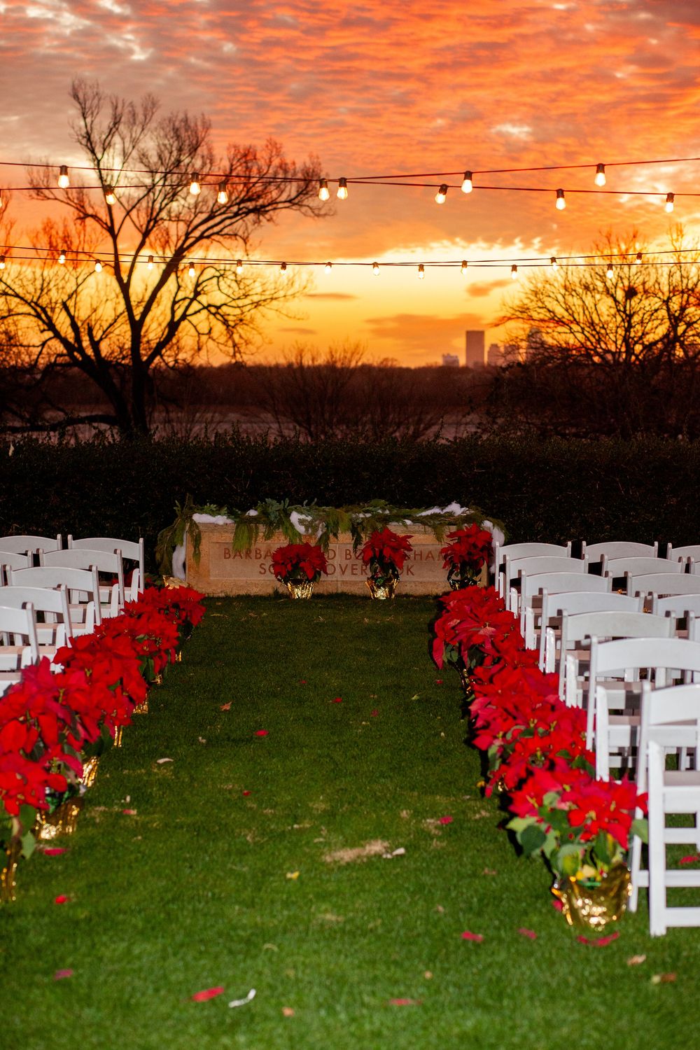 White chairs and red poinsettias line a grassy lawn at sunset with a dramatic orange sky and city skyline in background.