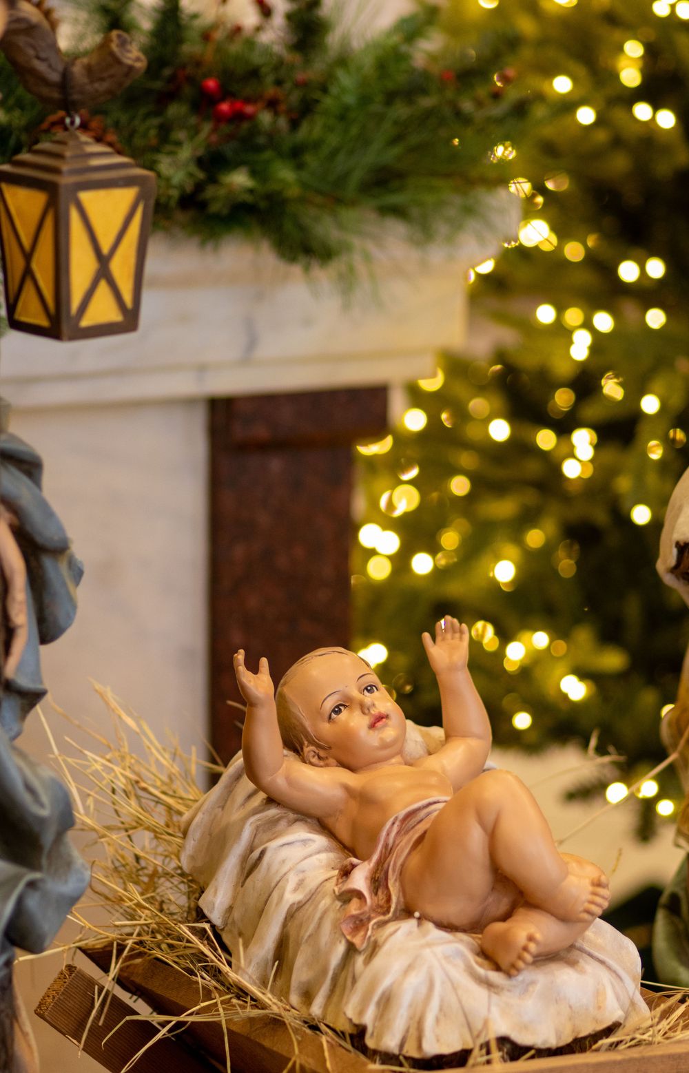Baby Jesus figurine in manger with Christmas tree lights and lantern decoration in background.