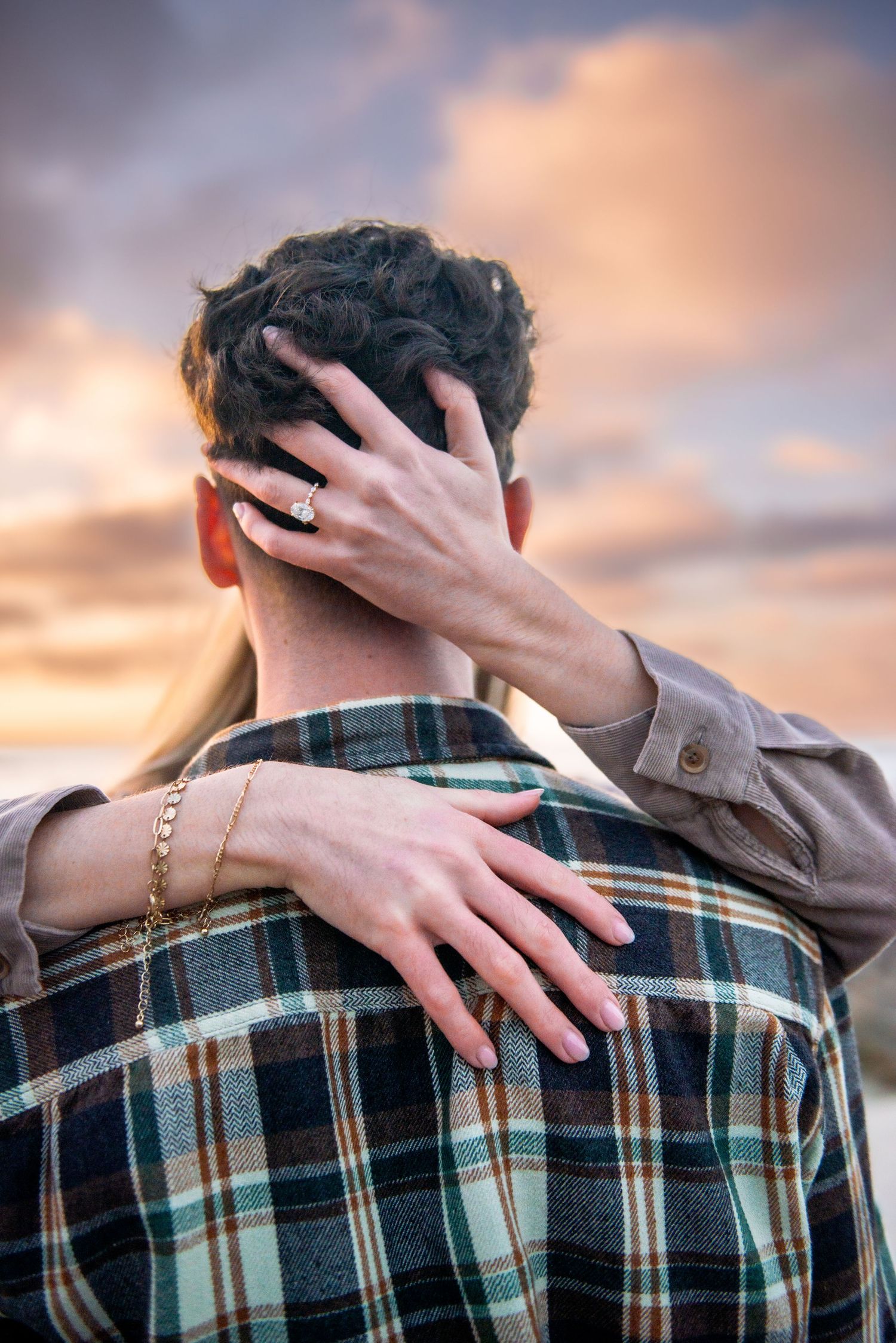 A hand reaches across to embrace someone wearing a plaid shirt against a sunset sky backdrop.