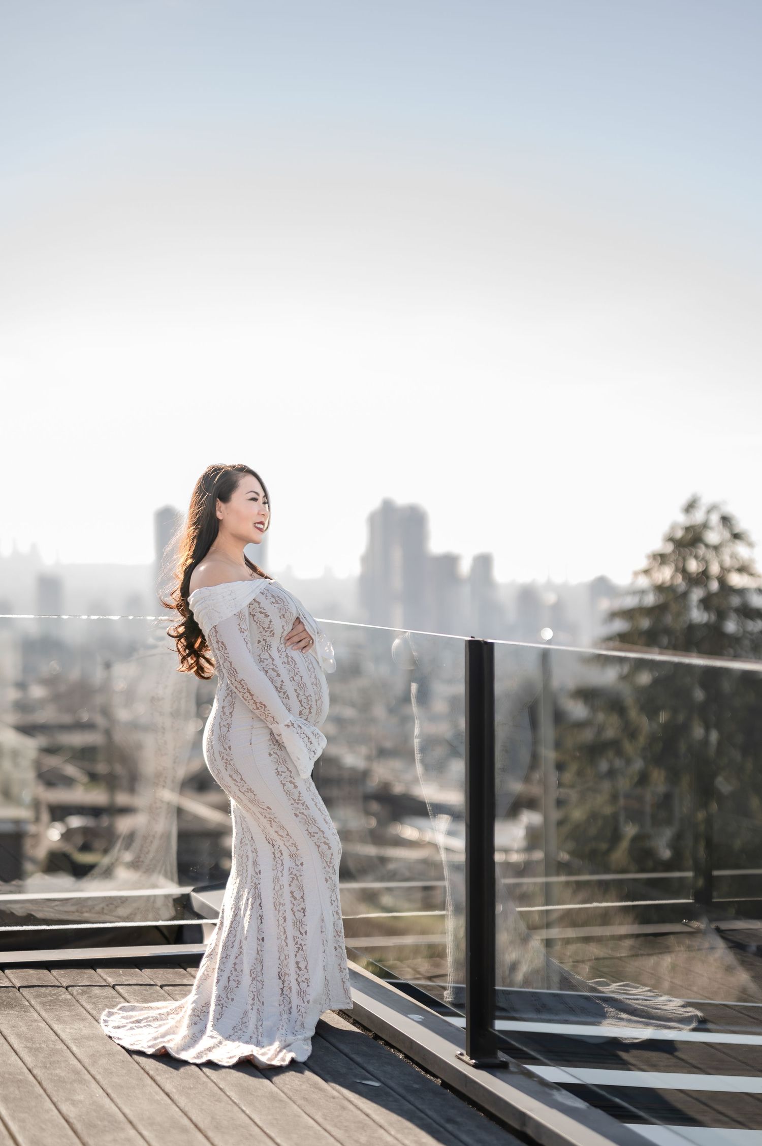A woman in a white lace dress poses elegantly near sheer curtains in a bright interior setting.