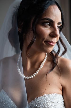 A bride wearing a pearl necklace and strapless beaded wedding dress poses in soft window light.