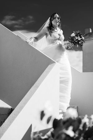 A bride in a fitted white wedding dress poses dramatically on white architectural stairs holding a bouquet in black and white.