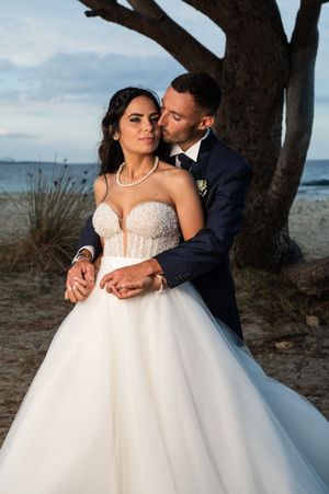 A romantic wedding photoshoot at sunset shows a couple embracing by a tree near the beach in elegant formal attire.