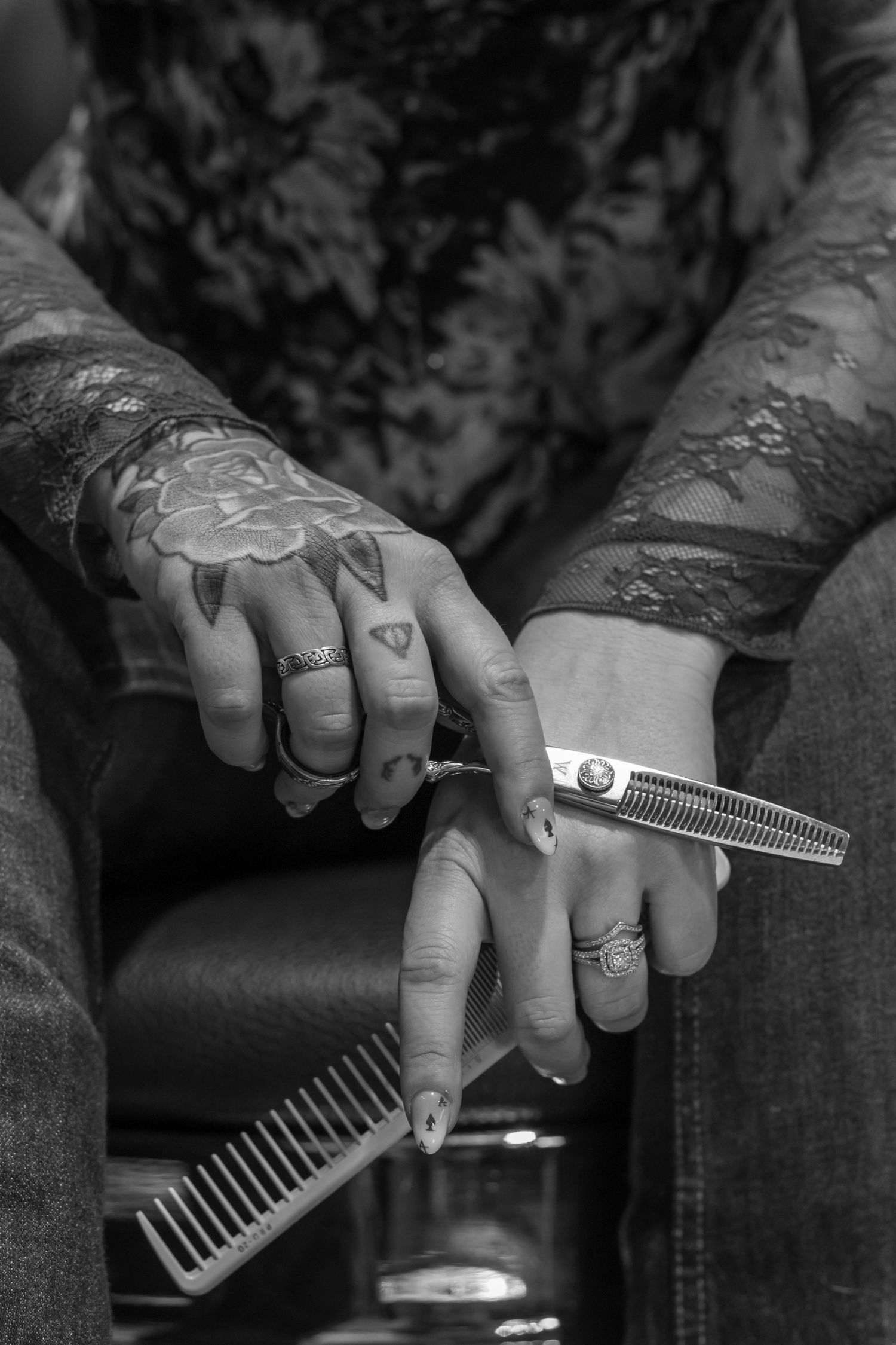 Black and white close-up of tattooed hands holding barber scissors and comb in a salon setting.