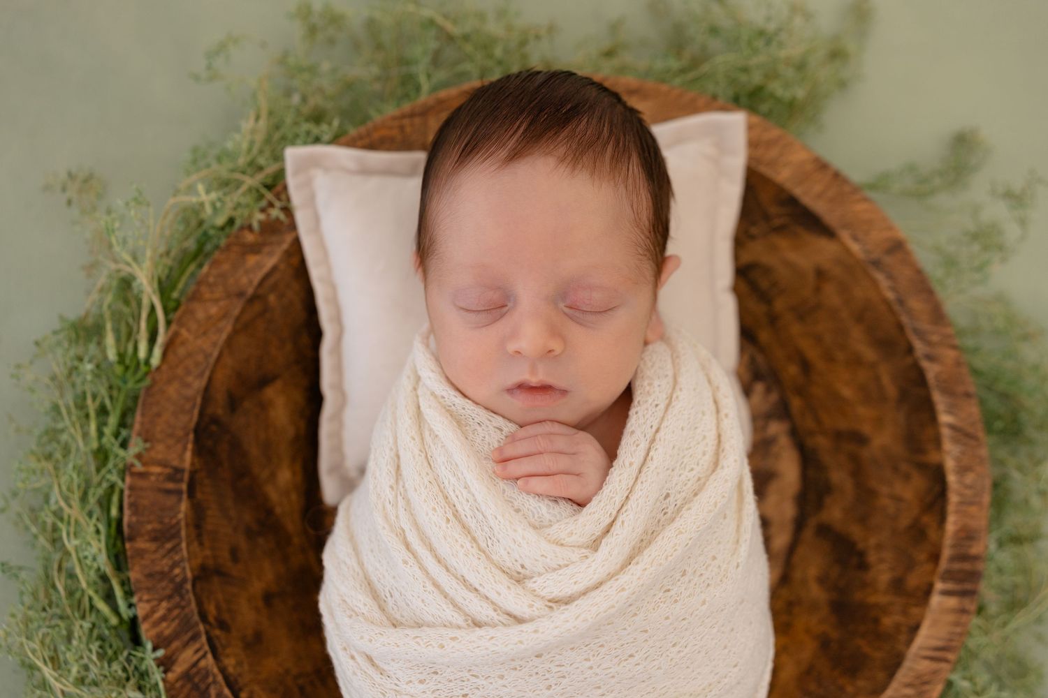 A sleeping newborn wrapped in a cream-colored blanket lies in a rustic wooden bowl with green foliage accents.