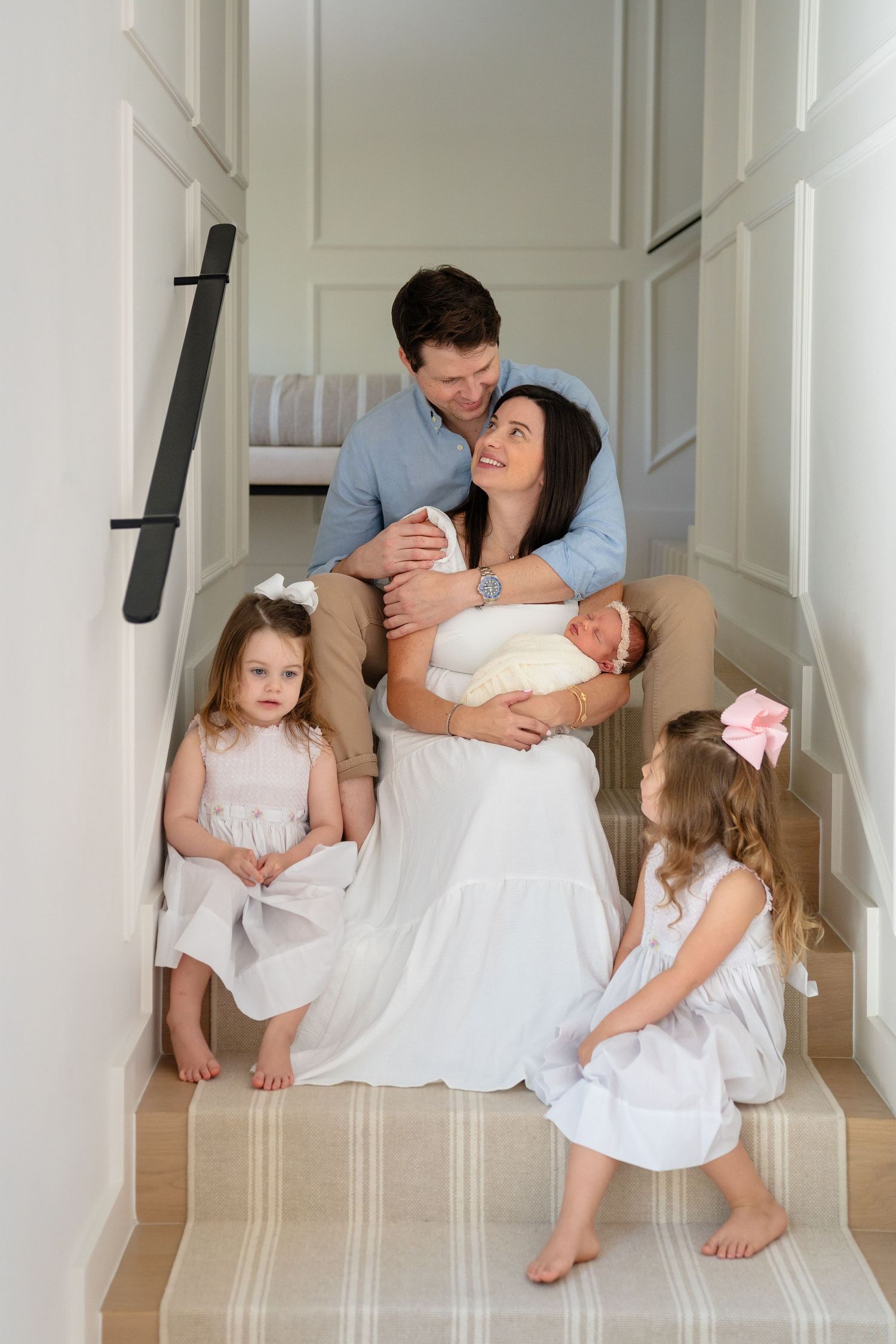 Family sitting together on carpeted stairs in white hallway wearing coordinated white and neutral outfits.