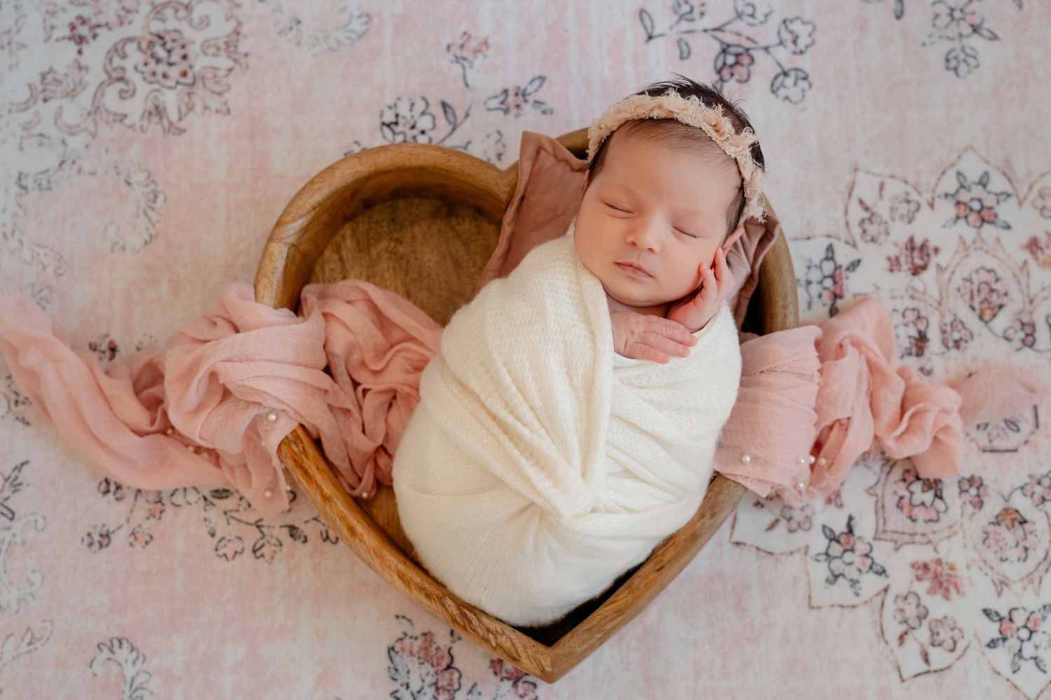 Newborn baby swaddled in white blanket lying on pink floral backdrop during professional photo session.