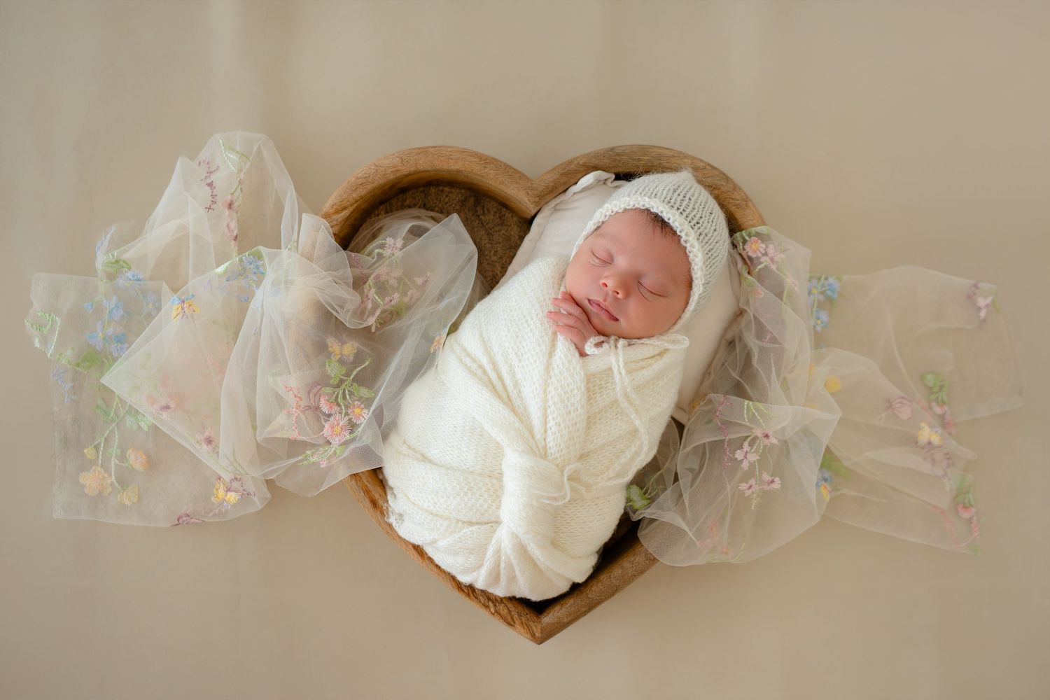 A sleeping newborn baby swaddled in white lies in a heart-shaped basket with delicate floral tulle decorations.
