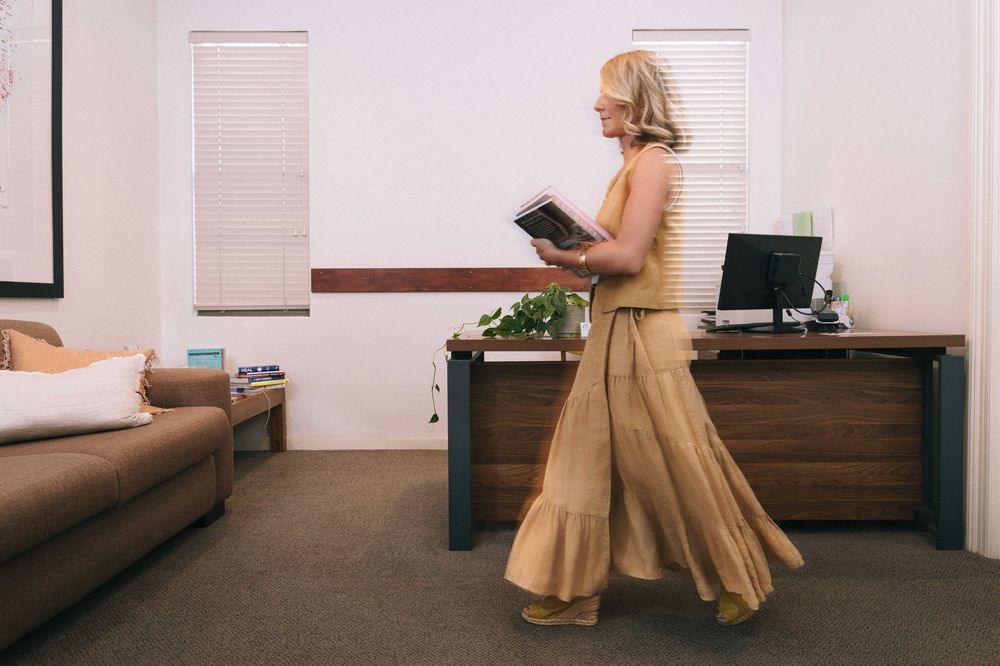 A person in a flowing tan dress walks through an office space while looking at a tablet device.