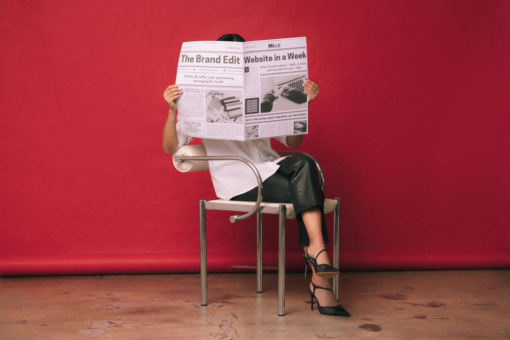 Woman in business attire seated on chair reading newspaper against vibrant red wall.