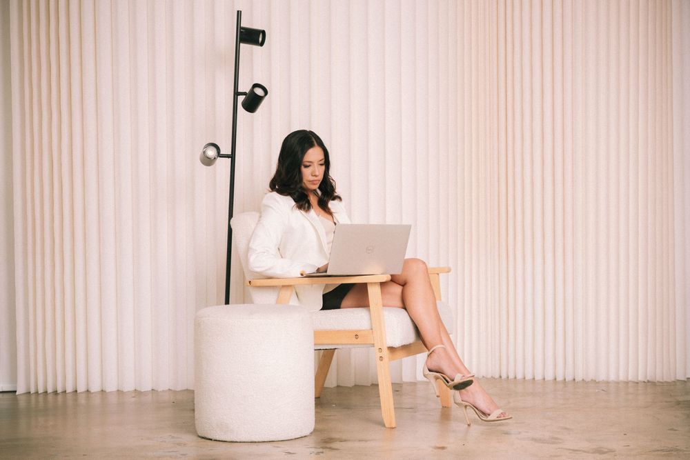 Woman in white dress sits elegantly on modern chair against beige curtained wall.