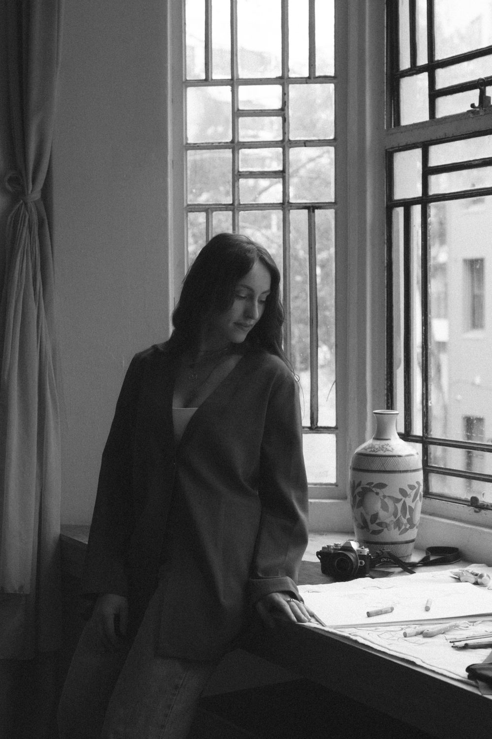 A black and white photograph of someone standing by a window with a vase and desk, captured in a contemplative moment.