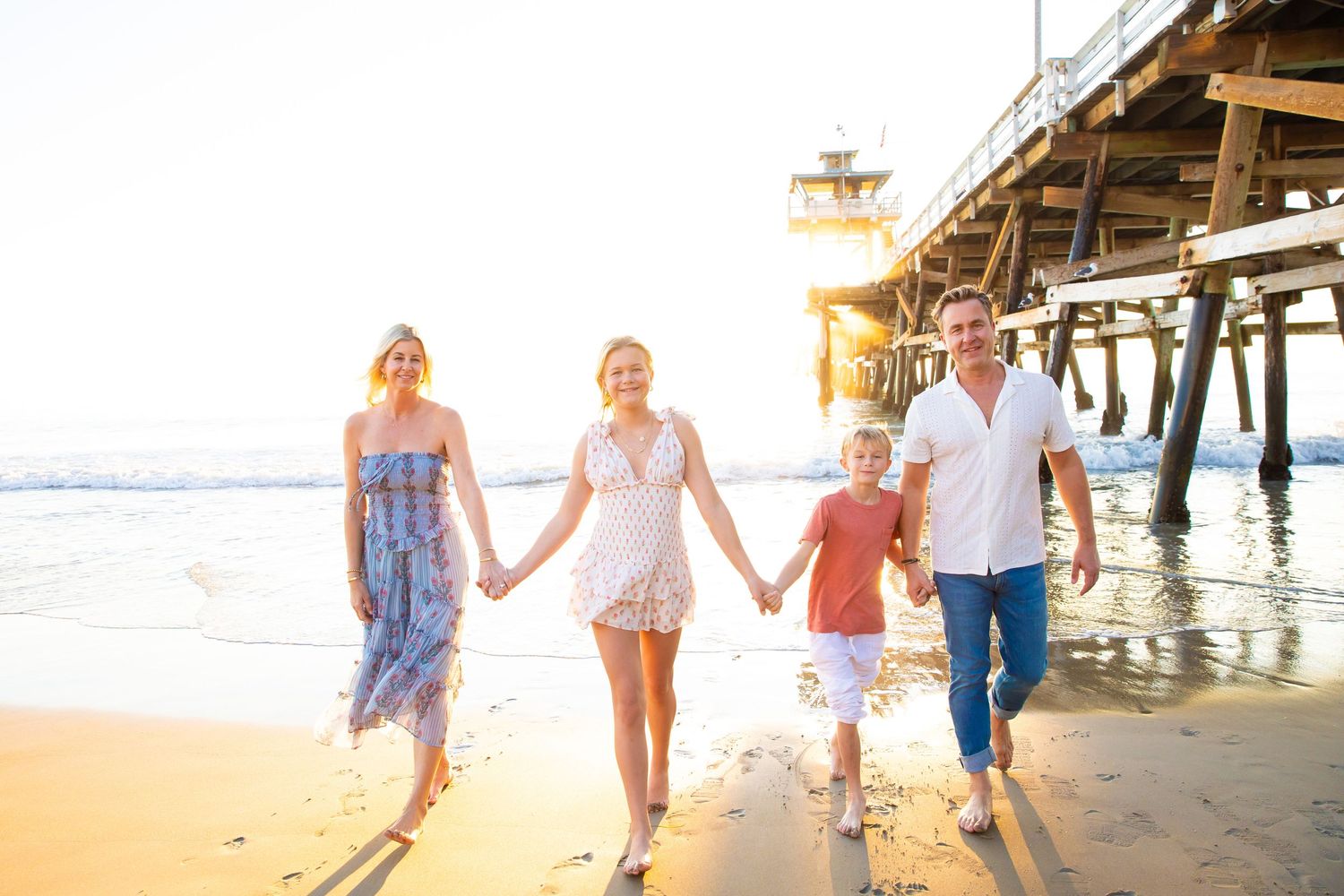 Family walking together holding hands on sandy beach at sunset near wooden pier.