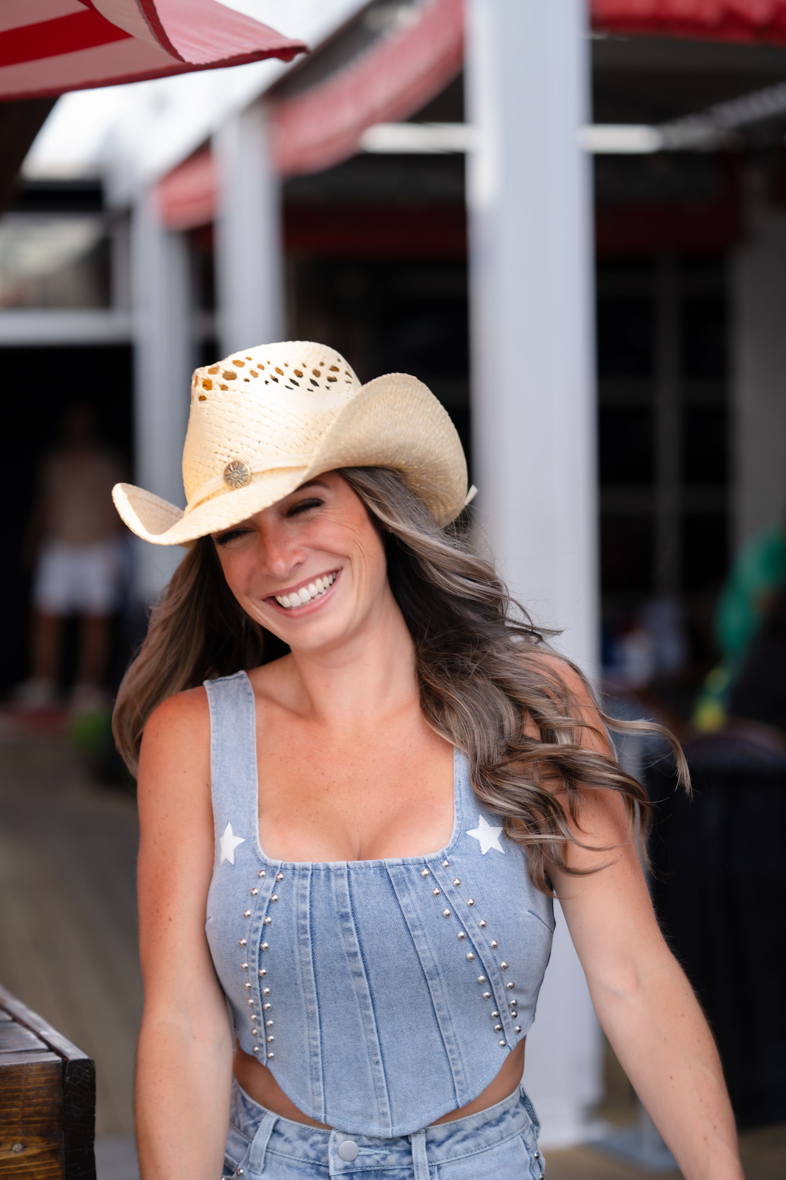 Woman in denim overalls and straw cowboy hat walking along wooden boardwalk under red awning.