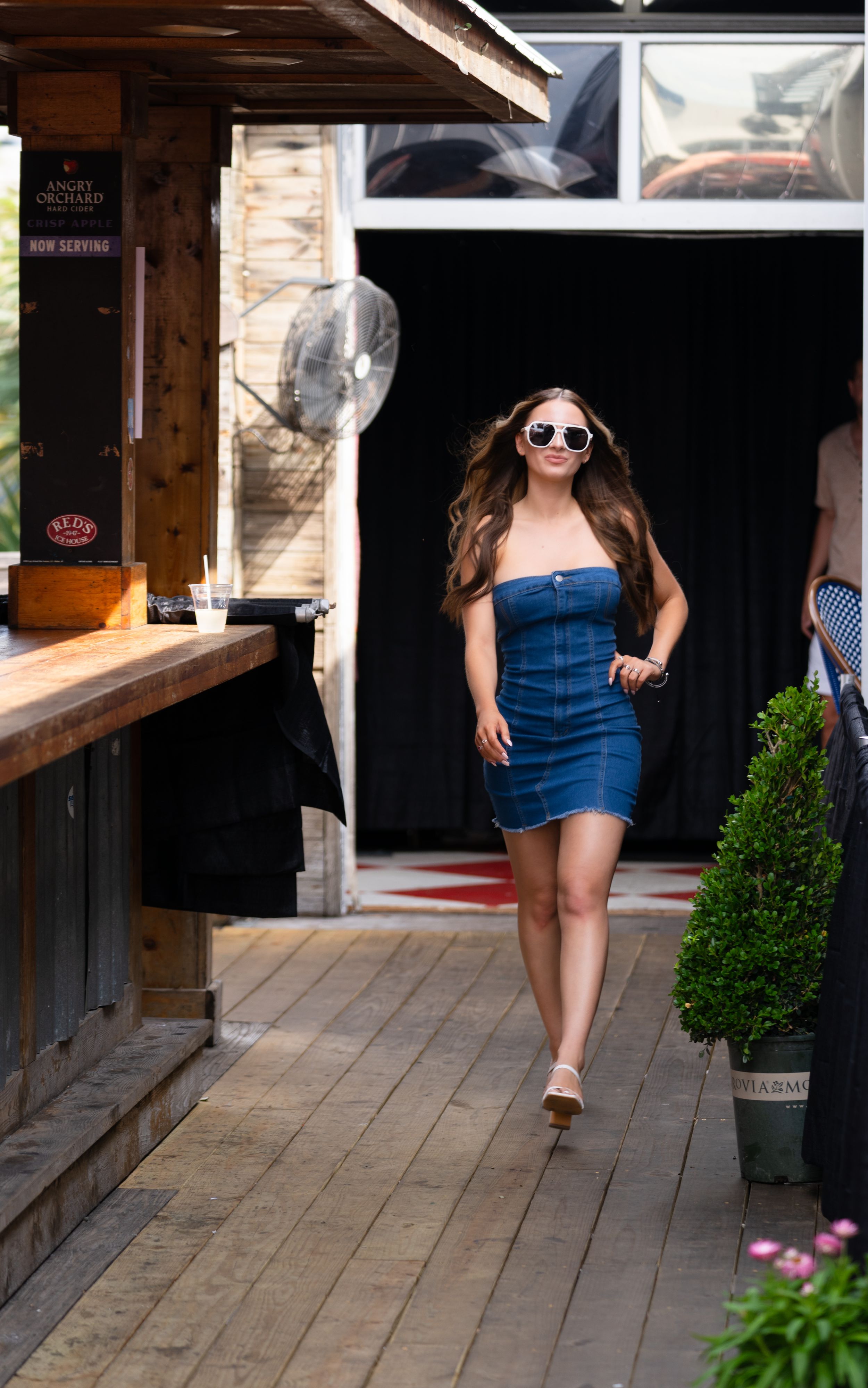 A person in a denim mini dress and sunglasses walks along a wooden deck with potted plants and outdoor seating.
