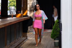 A person wearing a pink bikini walks along a wooden boardwalk near tropical plants and buildings.
