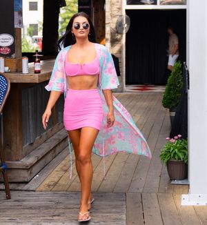 Woman in pink dress and floral cover-up walks along wooden boardwalk on sunny summer day.