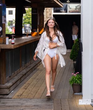 Woman in white swimsuit and sheer cover-up walks along wooden boardwalk near restaurant.