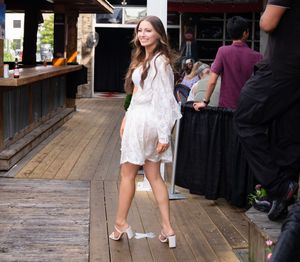 A person in a white lace dress and heels poses on a wooden deck near an outdoor bar area.