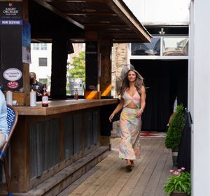 Woman in floral dress walks along covered wooden walkway outside retail shops.