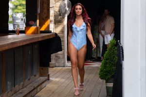 Woman in blue one-piece swimsuit poses on wooden deck with coastal decor in background.