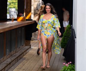 Woman in tropical print outfit walking on wooden deck with greenery and white pillars.