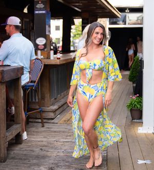 Woman in yellow floral coverup and white swimsuit poses on rustic wooden deck near beach bar.