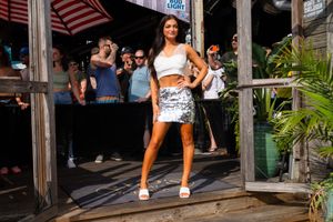 Woman in crop top and metallic mini skirt poses on outdoor deck with crowd in background.