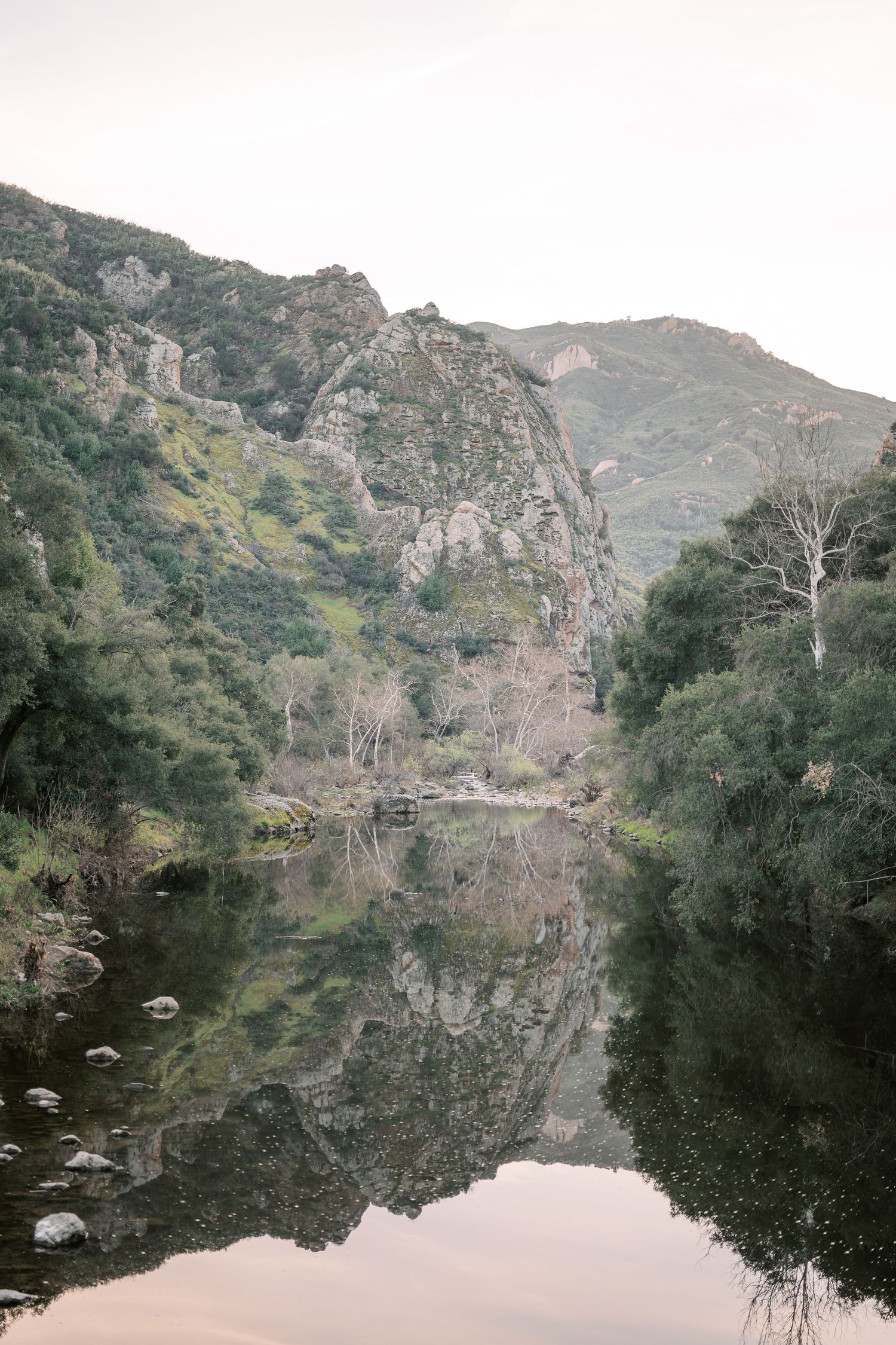 Scenic mountain landscape reflected in calm lake waters with rocky cliffs and trees along the shoreline.