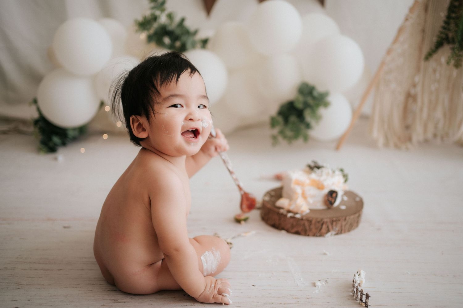 Baby sitting on white backdrop during cake smash photo session with white balloons and greenery decorations.