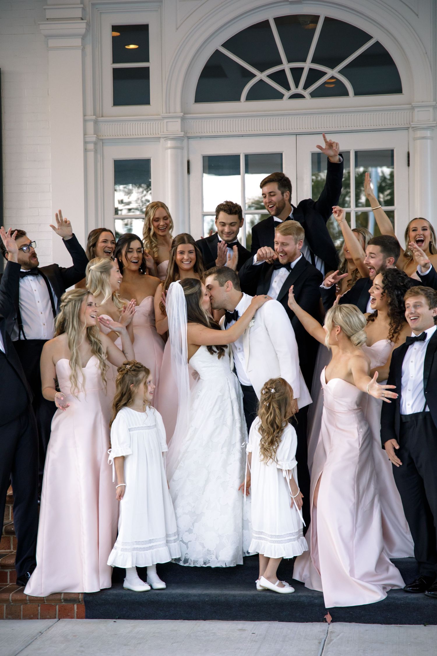 Joyful wedding party celebrates with raised arms in front of white colonial architecture.