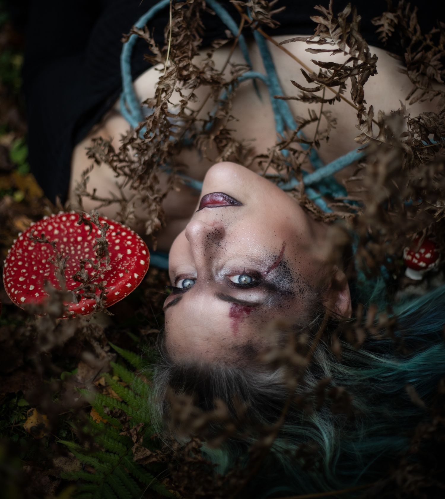 A dreamy portrait shows a person lying in dried foliage with blue hair and red berries in an artistic composition.