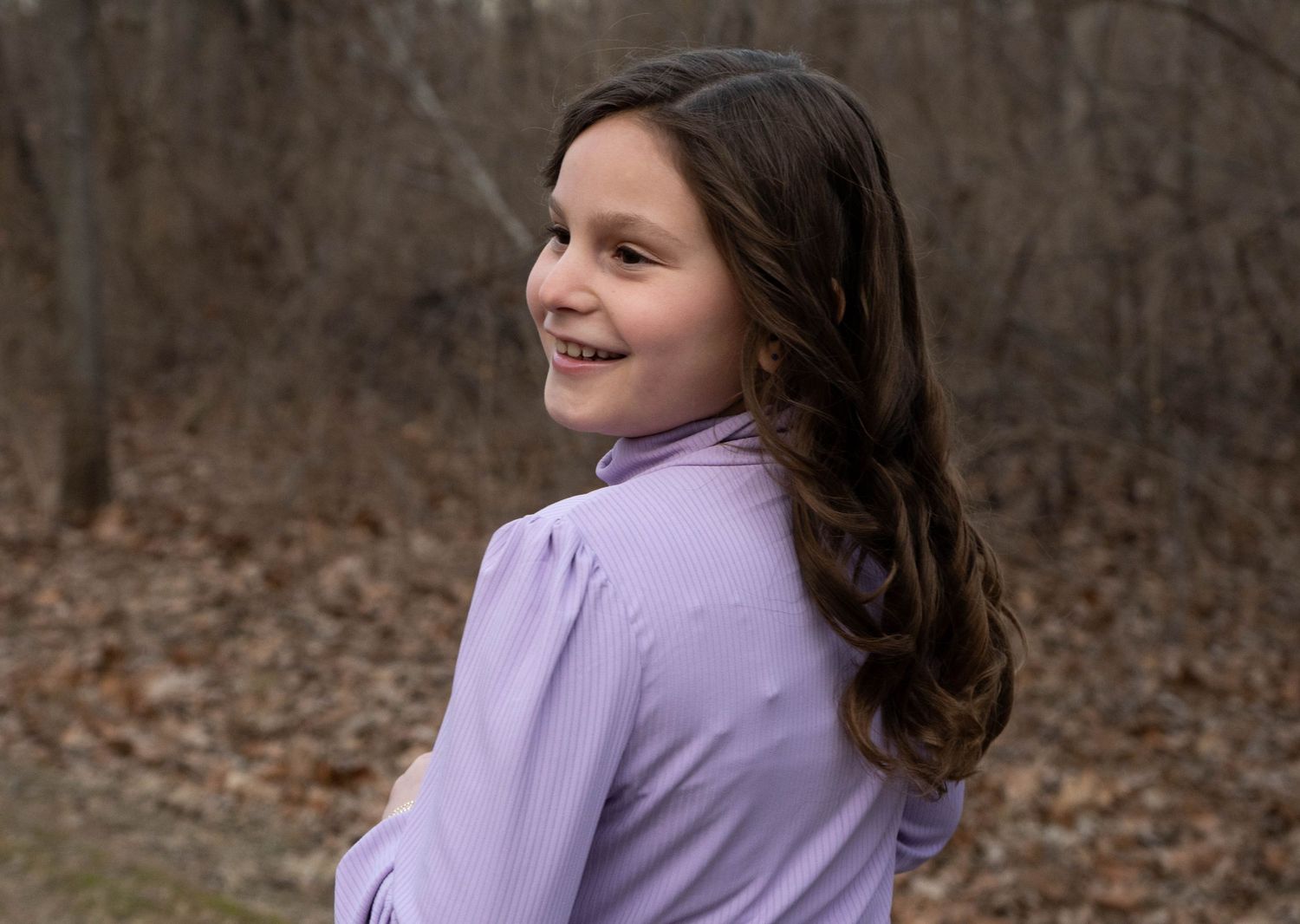Young girl with long brown hair smiling over her shoulder outdoors in a purple turtleneck in bare winter woods.