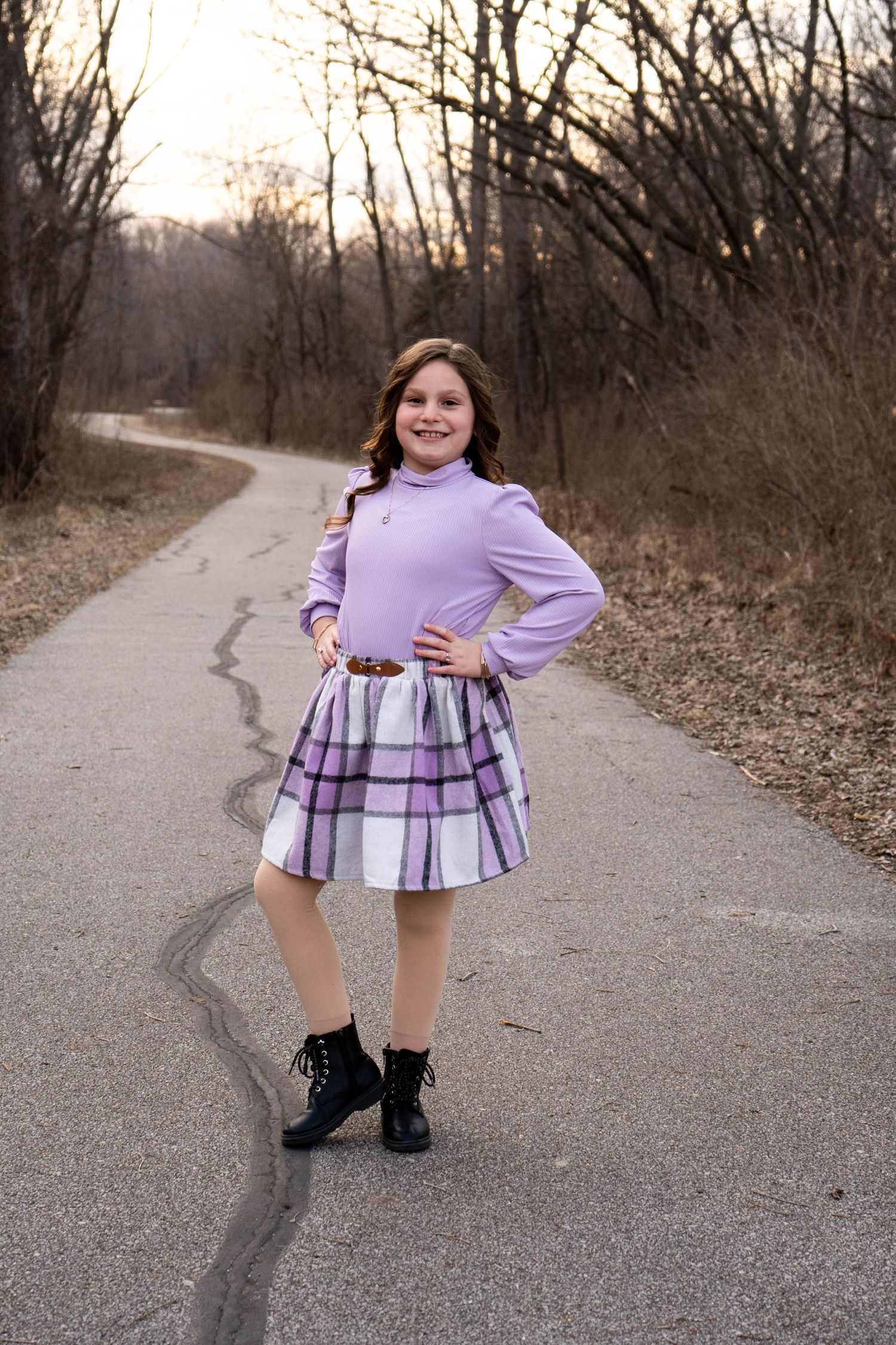 Confident young girl posing on a winding path in a purple turtleneck and plaid skirt with black lace-up boots.