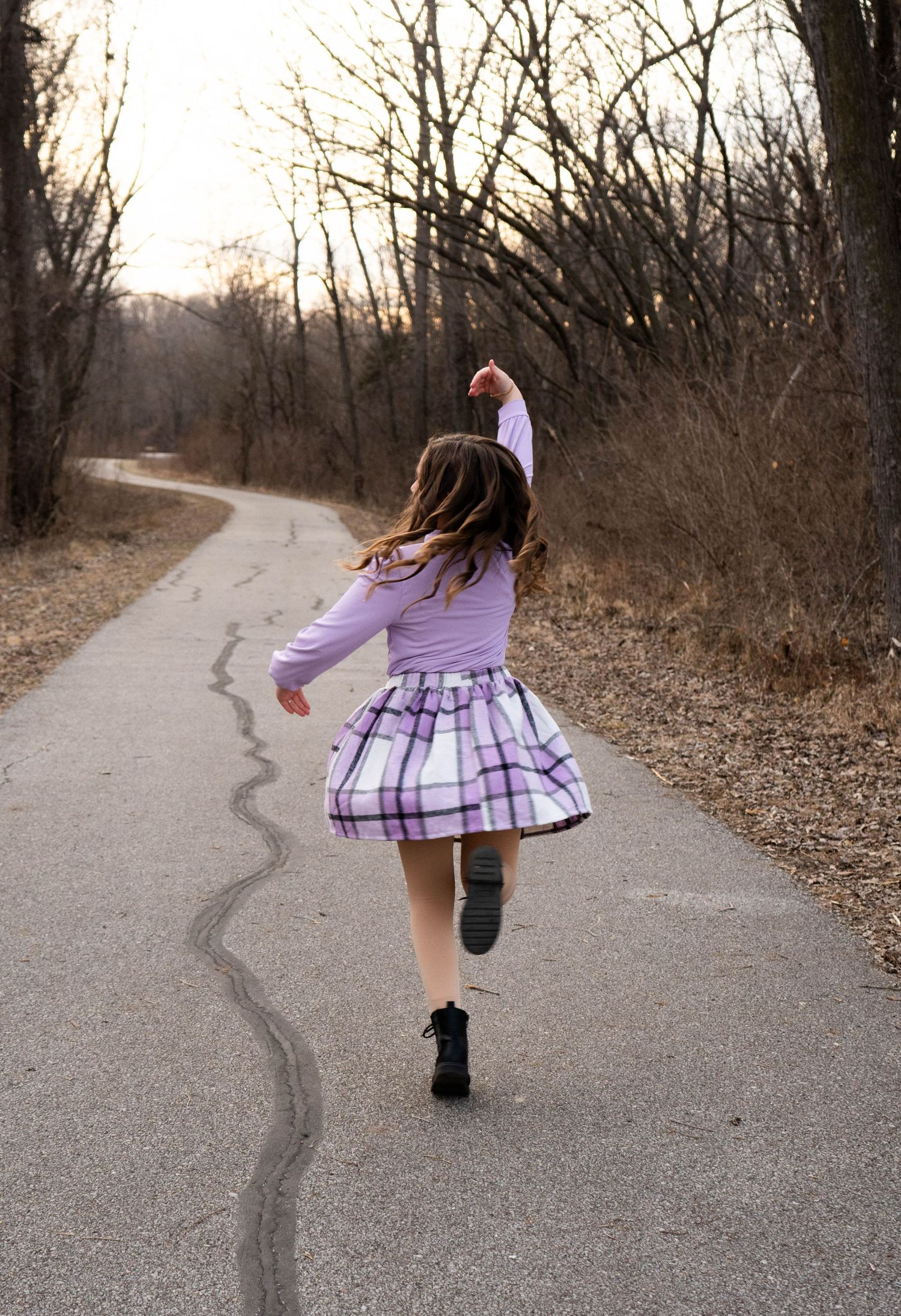 Young girl in purple plaid skirt twirling on winding park path at dusk.
