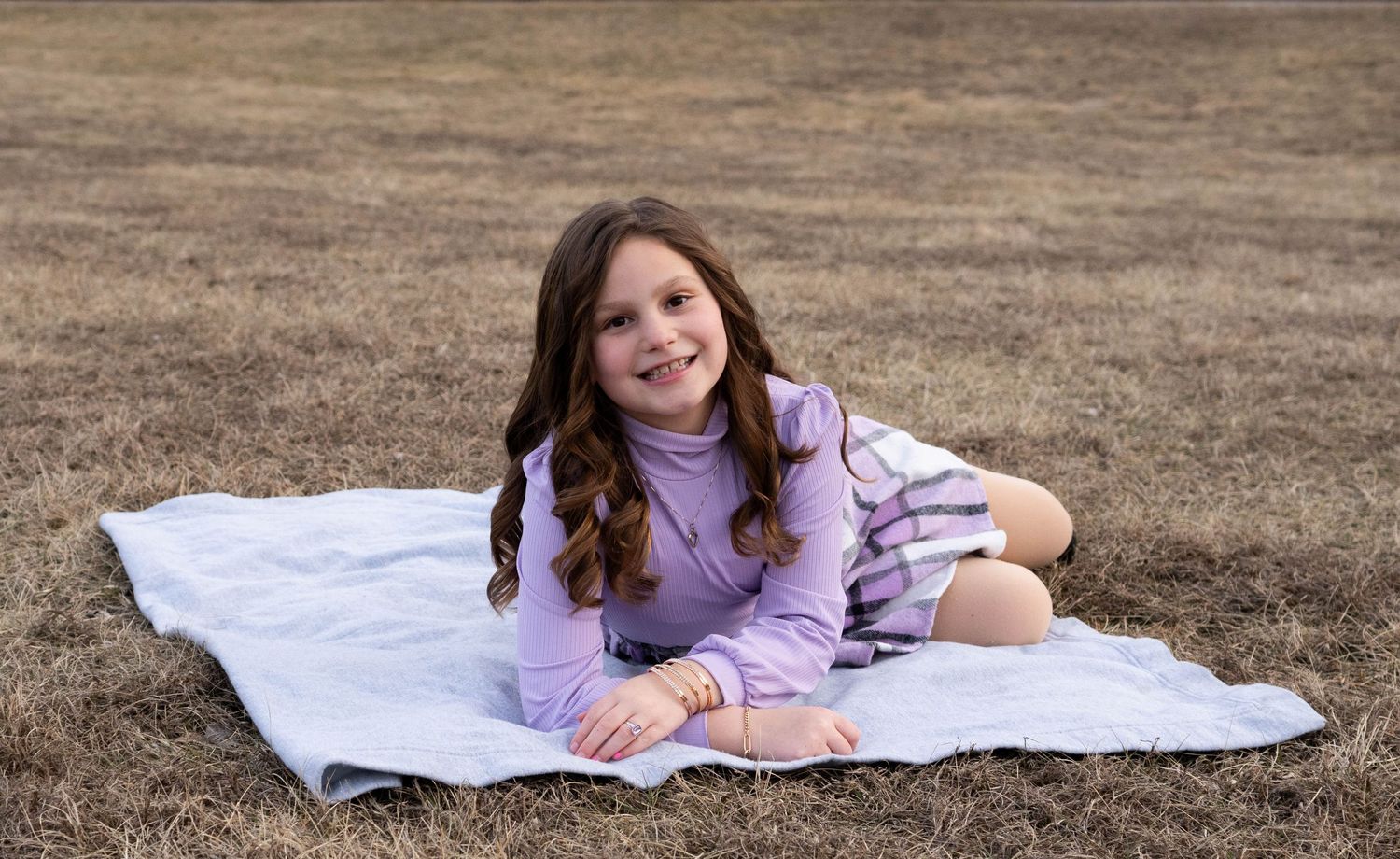 Cheerful girl sitting on white blanket in brown grass field smiling.