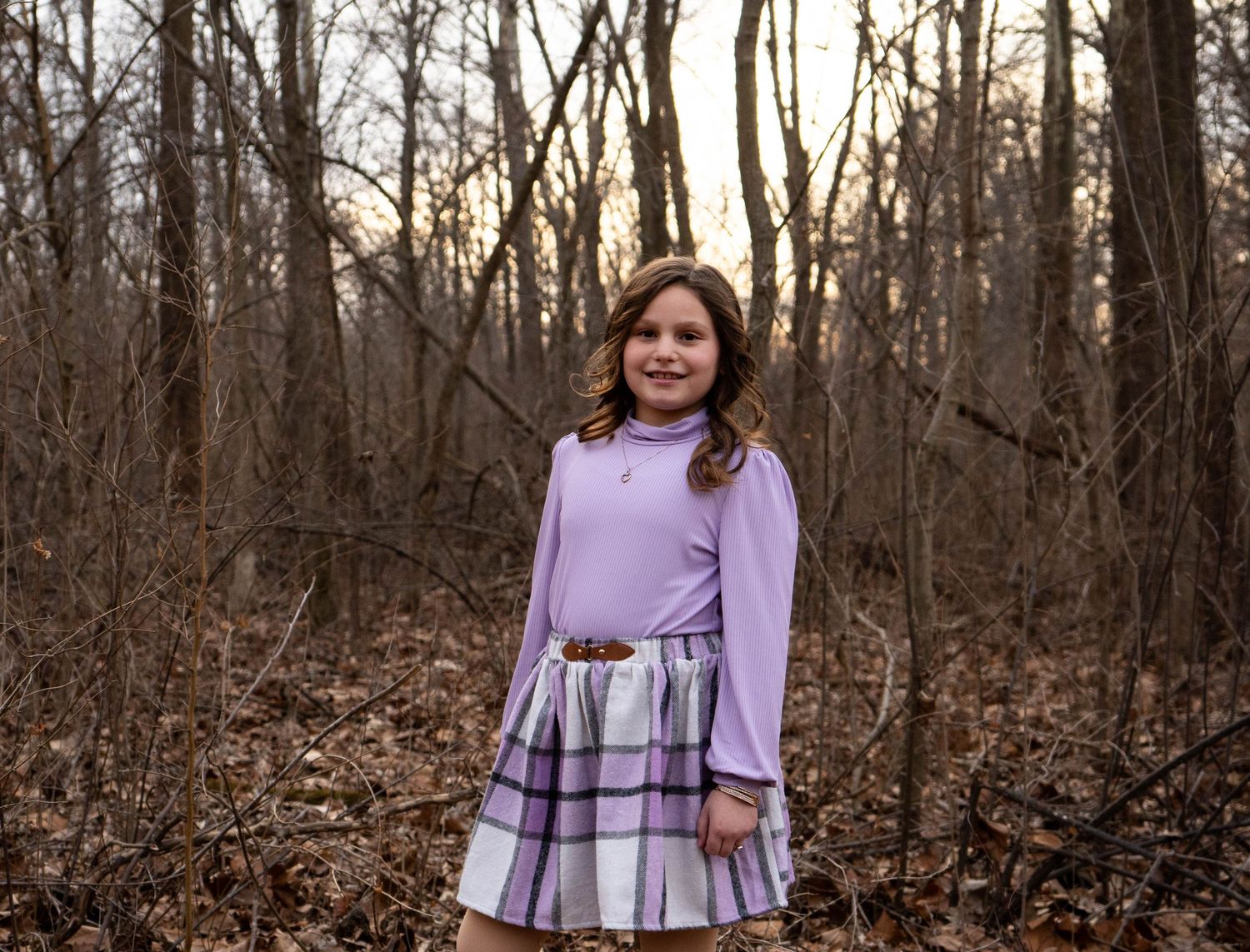 Young girl standing in bare winter woods wearing a purple turtleneck and plaid skirt with black ankle boots.