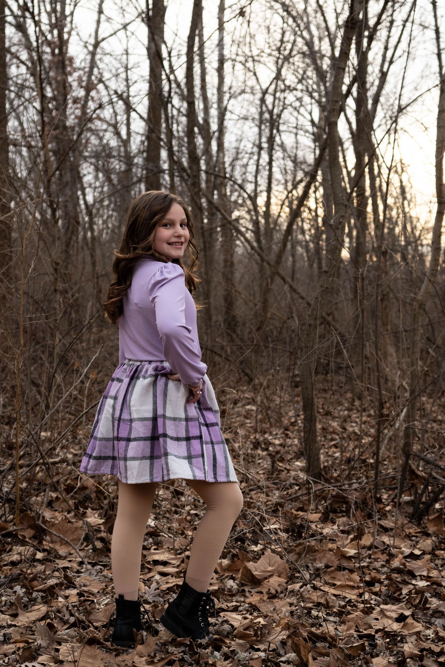 Young girl twirling on a leaf-covered path in bare winter woods, wearing purple turtleneck and plaid skirt.