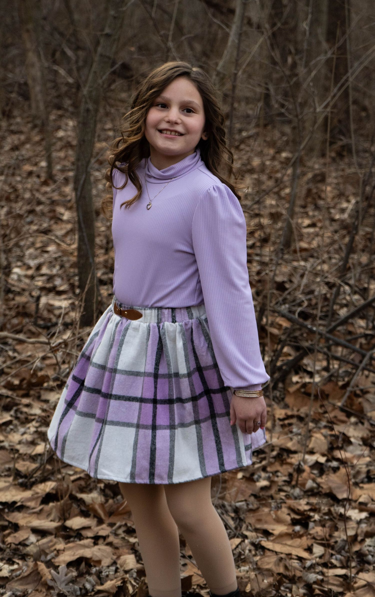 Smiling young girl standing in leaf-covered woods in a purple turtleneck and plaid skirt with black boots.